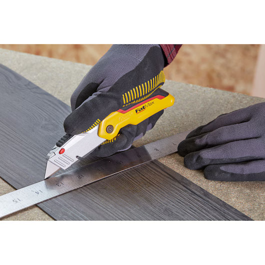 A yellow and silver Festool utility knife being used by a person wearing dark gray work gloves to cut a dark wood strip with a metal ruler on a workbench. STANLEY® FATMAX® Retractable Folding Utility Knife