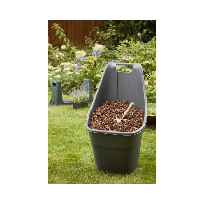 A dark gray plastic mulch bag filled with brown wood chips and a small hand rake, set on a green lawn in a garden with plants and a wooden fence in the background.