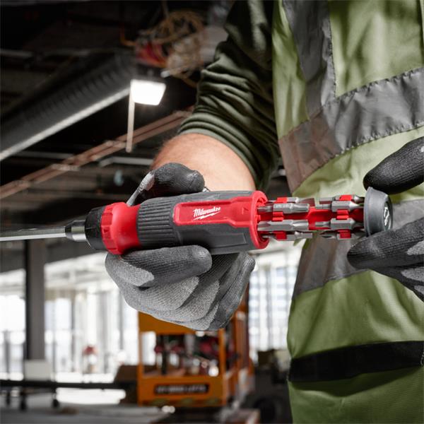 A close-up of a person wearing a high-visibility safety vest and gloves, using a Milwaukee cordless screwdriver with multiple bits attached in an industrial workshop setting.