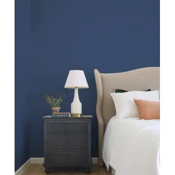 A styled bedroom corner featuring a deep navy blue wall, a dark charcoal nightstand with a white table lamp and small vase, and a bed with white linens and a peach accent pillow.