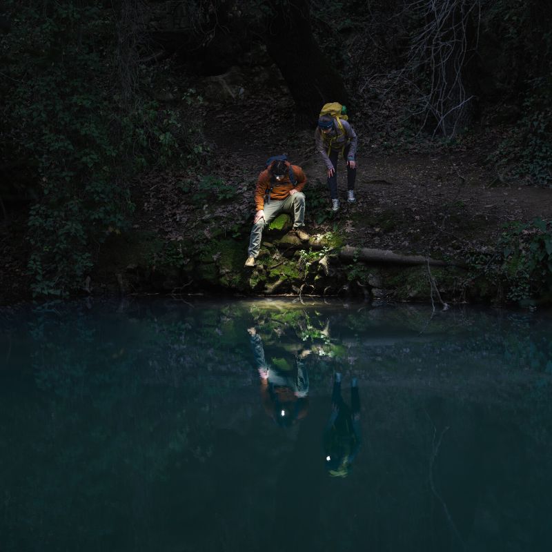 Two hikers with backpacks are seen from above on a dark forest path beside a calm dark blue-green river, with their reflections visible in the water.