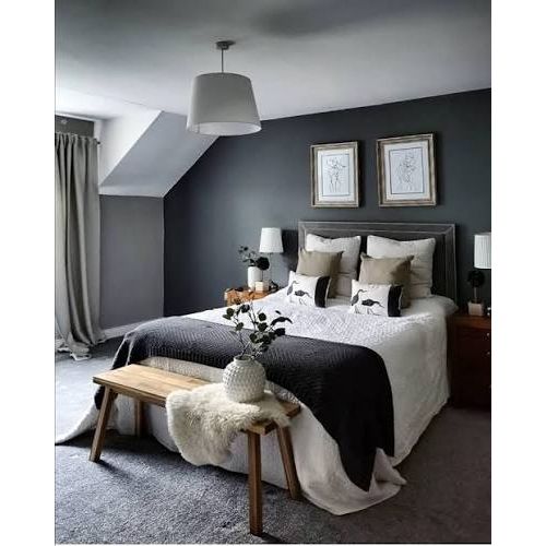 A styled bedroom interior featuring a bed dressed in white and black bedding, set against a dark gray wall with two framed prints. A wooden bench with a white faux-fur throw sits at the foot of the bed, and a gray rug covers the floor.