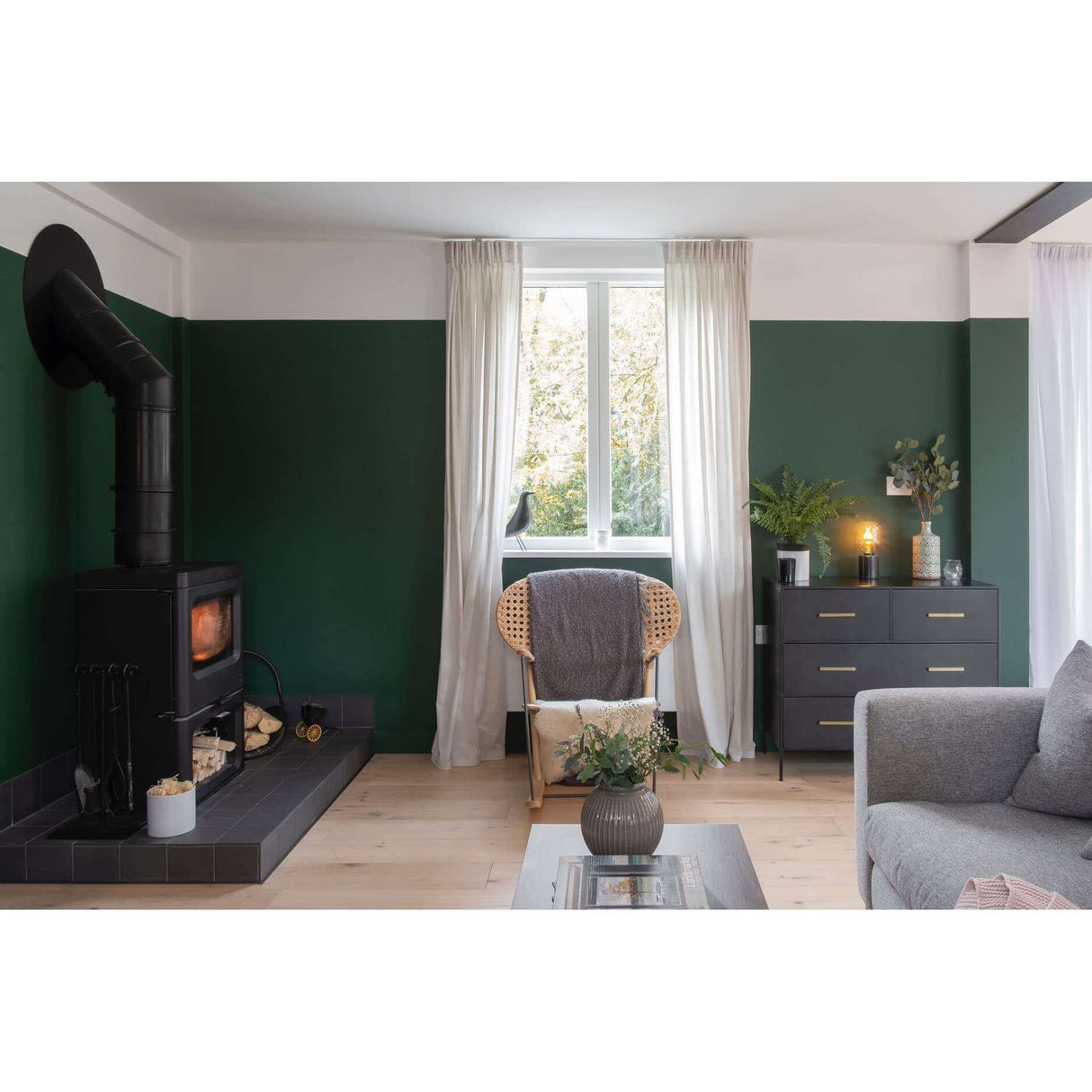 A styled living room interior with dark green walls, white trim, a black wood-burning stove with a tall black chimney pipe, sheer white curtains, a woven armchair with a gray throw, a dark gray dresser, and part of a gray sofa visible.