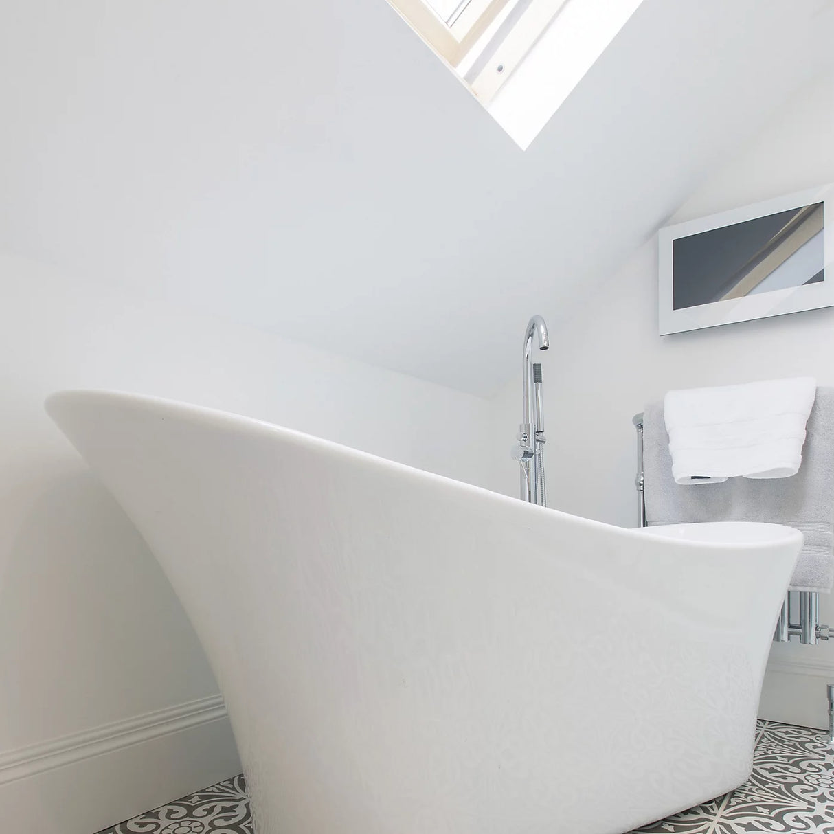 A modern white freestanding bathtub in a bright, minimalist bathroom with white walls, a patterned tile floor, and a skylight.