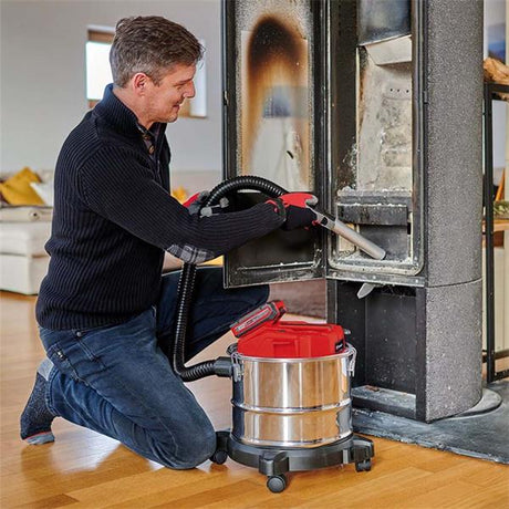 A man kneels on a wooden floor using a cordless floor vacuum cleaner with a red lid and a flexible hose to clean the interior of a tall fireplace or stove.