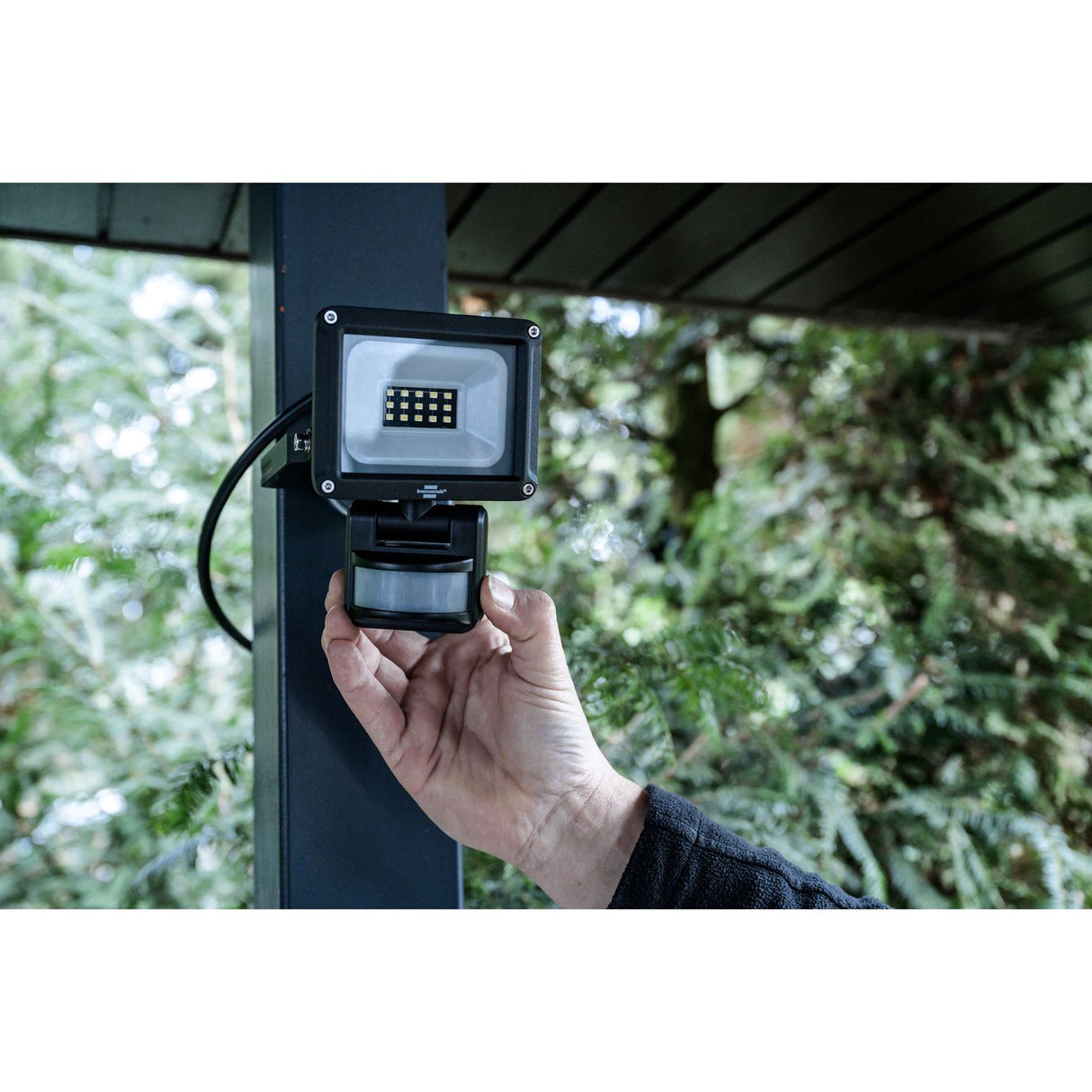 A black outdoor LED floodlight with an attached motion sensor, mounted on a dark post with a hand holding the sensor in front of a blurred green garden background. Brennenstuhl JARO 1060 P LED Spotlight with Motion Detector | 10W | 1150 Lumen | IP65