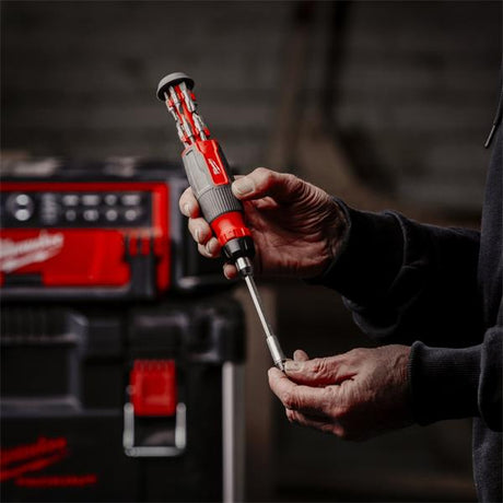 A person's hands holding a Milwaukee cordless screwdriver with a multi-bit driver bit attachment, with a Milwaukee tool case blurred in the background.