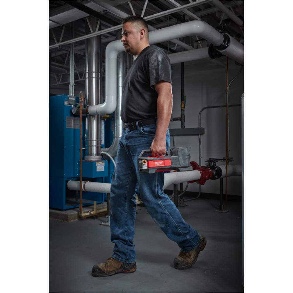 A man in a workshop or utility room carrying a red and gray cordless tool in his hand, wearing a dark gray t-shirt, blue jeans, and brown work boots. Milwaukee M18BTP-0 M18 Transfer Pump | Bare Unit