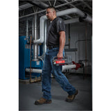 A man in a workshop or utility room carrying a red and gray cordless tool in his hand, wearing a dark gray t-shirt, blue jeans, and brown work boots. Milwaukee M18BTP-0 M18 Transfer Pump | Bare Unit