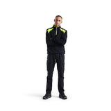 A man wearing a black high-visibility work jacket with neon yellow-green shoulder panels and matching black cargo pants, standing against a solid white background.