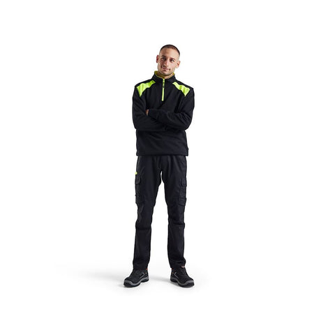 A man wearing a black high-visibility work jacket with neon yellow-green shoulder panels and matching black cargo pants, standing against a solid white background.