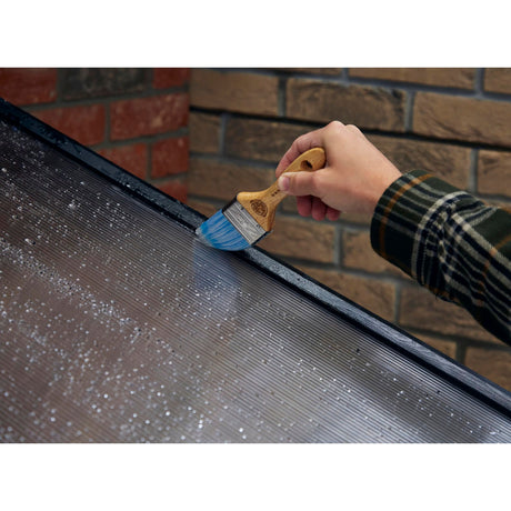 A close-up of a hand holding a paint brush with blue bristles and a light wood handle, applying paint to the edge of a dark metal solar panel array. A brick wall is visible in the background.
