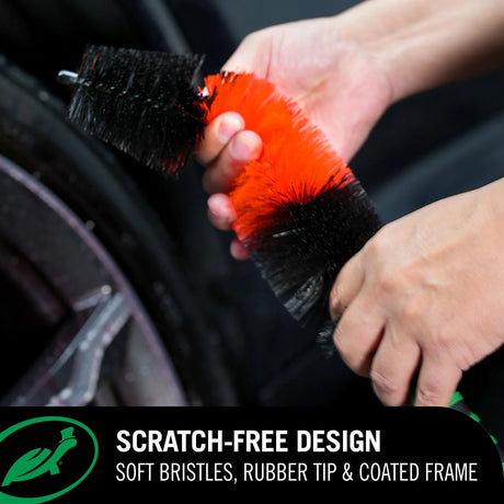Person holding a car cleaning brush with red bristles and black handle, with a car wheel in the background.