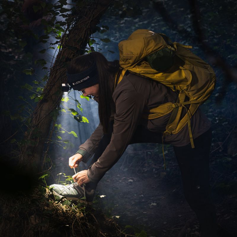 A woman hiking in a dark forest at night, wearing a large yellow backpack, a brown jacket, and a headlamp while tying the strap of her hiking boot.