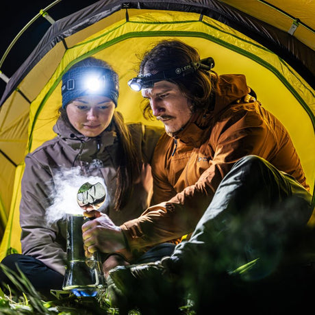 A man and a woman in outdoor gear sit inside a yellow tent at night, using headlamps to illuminate a glass smoking apparatus. The man wears an orange-brown jacket, and the woman wears a gray-brown jacket.