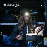 A woman wearing a LEDLENSER headlamp with a bright white light on, standing inside a vehicle or trailer next to a camping stove and pot. The LEDLENSER brand logo is visible in the top left corner.