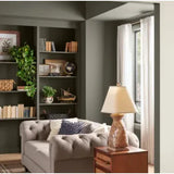 Prestige Boot Room
A styled interior room scene featuring a tufted light gray sofa with patterned throw pillows, a wooden side table with a lit table lamp, built-in dark green bookshelves with books and plants, and a window with sheer white curtains.