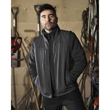 A man wearing a dark gray zip-up work jacket with a high collar, standing in a workshop with tools and equipment in the background.