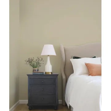 A styled bedroom corner featuring a dark charcoal gray three-drawer nightstand with a white table lamp and a small vase of flowers, next to a bed with white linens and a peach accent pillow against a warm beige wall.