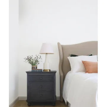 A styled bedroom corner featuring a dark charcoal gray three-drawer nightstand next to a bed with a beige upholstered headboard, white bedding, and a peach accent pillow.