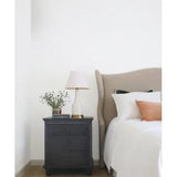 A styled bedroom corner featuring a dark charcoal gray three-drawer nightstand next to a bed with a beige upholstered headboard, white bedding, and a peach accent pillow.