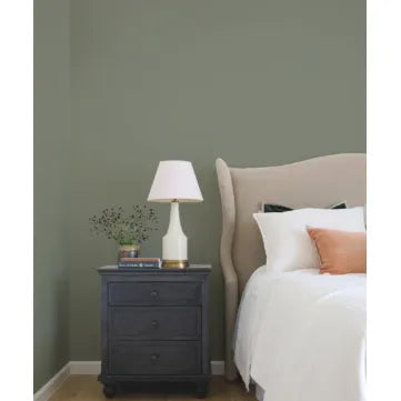 Bedroom with a nightstand, lamp, and bed with pillows against a green wall. Prestige Beau Paint