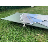 An outdoor green metal cot or folding lounger set on grass, with a blue and gray striped fringed blanket draped across it.