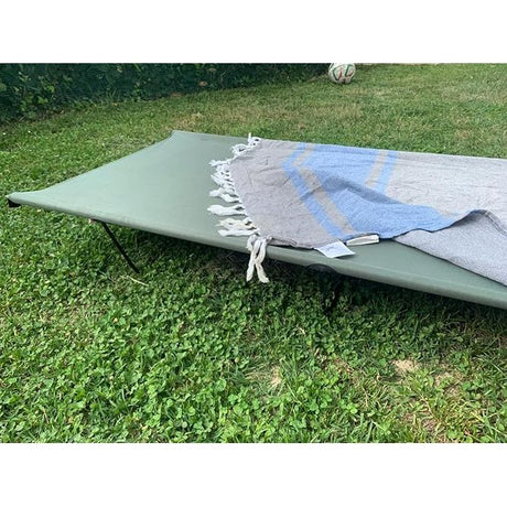 An outdoor green metal cot or folding lounger set on grass, with a blue and gray striped fringed blanket draped across it.
