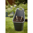 A gray plastic wheelbarrow filled with dark soil and two small hand tools, set on a green lawn in a garden with plants and a wooden fence in the background.