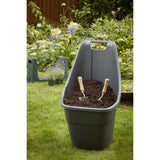 A gray plastic wheelbarrow filled with dark soil and two small hand tools, set on a green lawn in a garden with plants and a wooden fence in the background.