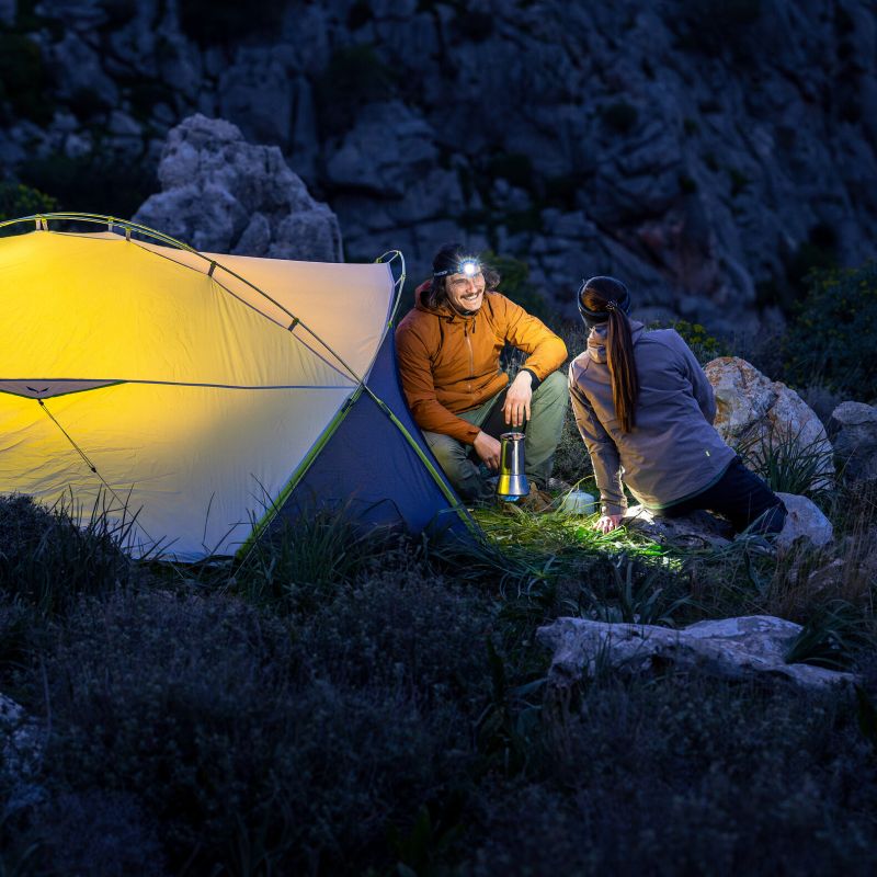 A couple camping at night beside a glowing yellow tent, with rocky mountain terrain in the background. One person wears an orange jacket and holds a lantern, while the other kneels nearby in the dark outdoor setting.