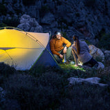 A couple camping at night beside a glowing yellow tent, with rocky mountain terrain in the background. One person wears an orange jacket and holds a lantern, while the other kneels nearby in the dark outdoor setting.