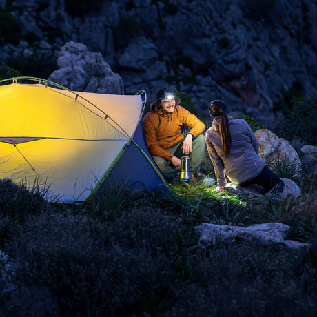A couple camping at night beside a glowing yellow tent, with rocky mountain terrain in the background. One person wears an orange jacket and holds a lantern, while the other kneels nearby in the dark outdoor setting.