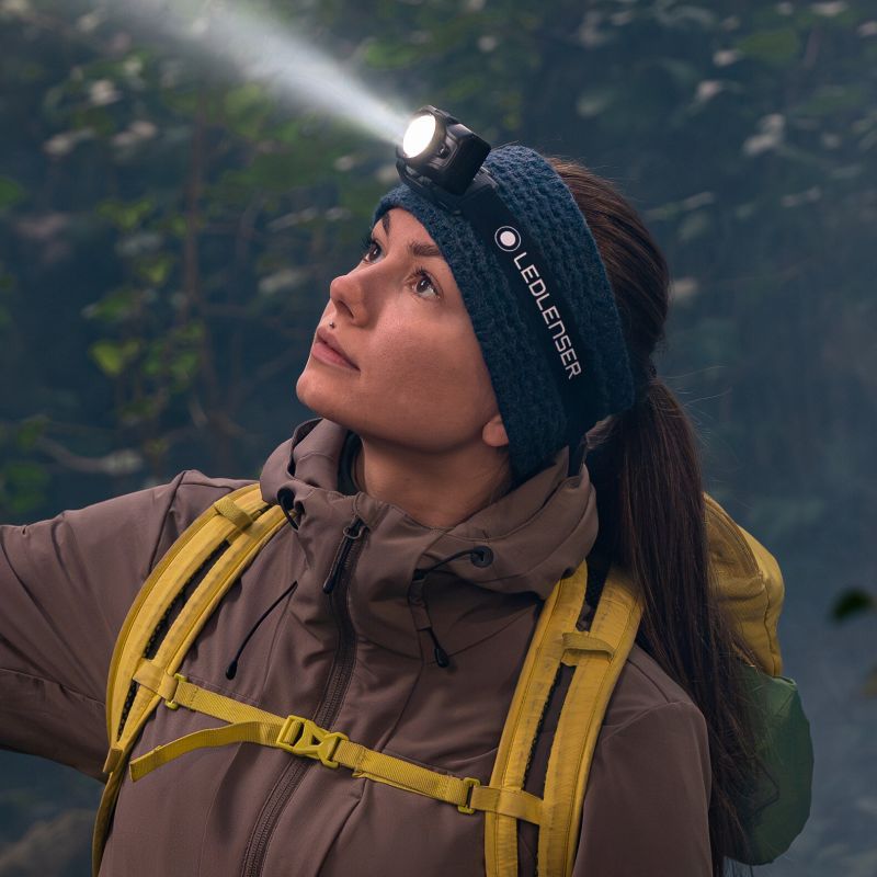A woman outdoors wearing a dark blue LED headlamp on a matching knit headband, with the beam of light shining upward. She is dressed in a brown jacket with yellow harness straps and a green backpack, set against a blurred forest background. Ledlenser Headlamp HF6R | 900 Lumen