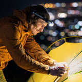 A man wearing a yellow waterproof jacket and a LED headlamp sets up a yellow tent at night, with blurred city lights in the background. The headlamp has the brand name 'LEGLEFTSER' visible.