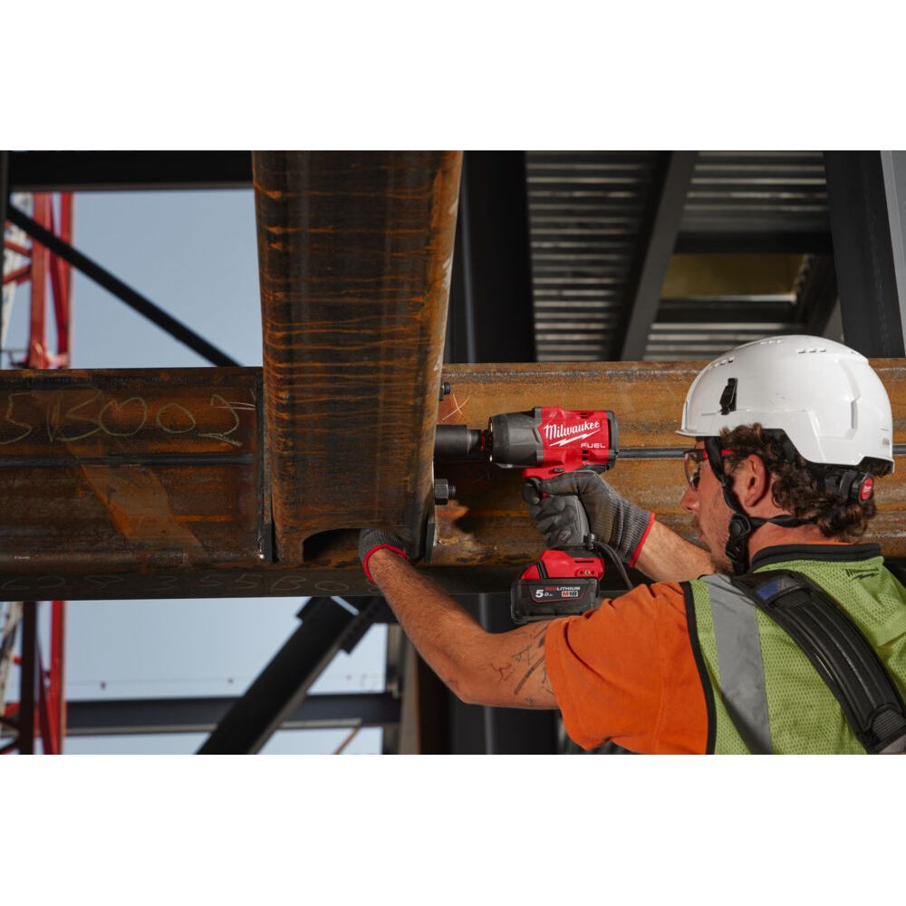 A construction worker wearing a white hard hat, orange shirt, and green safety vest uses a red and black Milwaukee cordless impact wrench to work on a large rusty steel beam in an industrial setting. 
Milwaukee M18 Fuel High Torque Impact Wrench With Friction Ring M18FHIW2F12-0