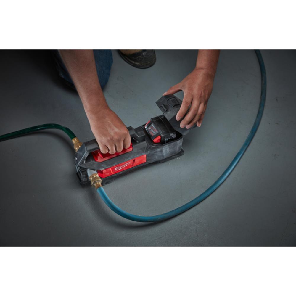 A person using a Milwaukee cordless nail gun with a blue air hose on a dark gray floor.