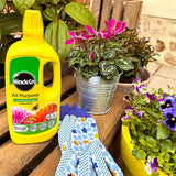 A bottle of Miracle-Gro All Purpose Flower Food fertilizer sits on a wooden table next to gardening gloves and potted plants in an outdoor garden setting.