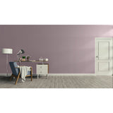 A minimalist room scene with a dusty mauve-pink wall, light wood floor, a white door on the right, and a home office setup on the left featuring a blue armchair, desk, lamp, and side table.