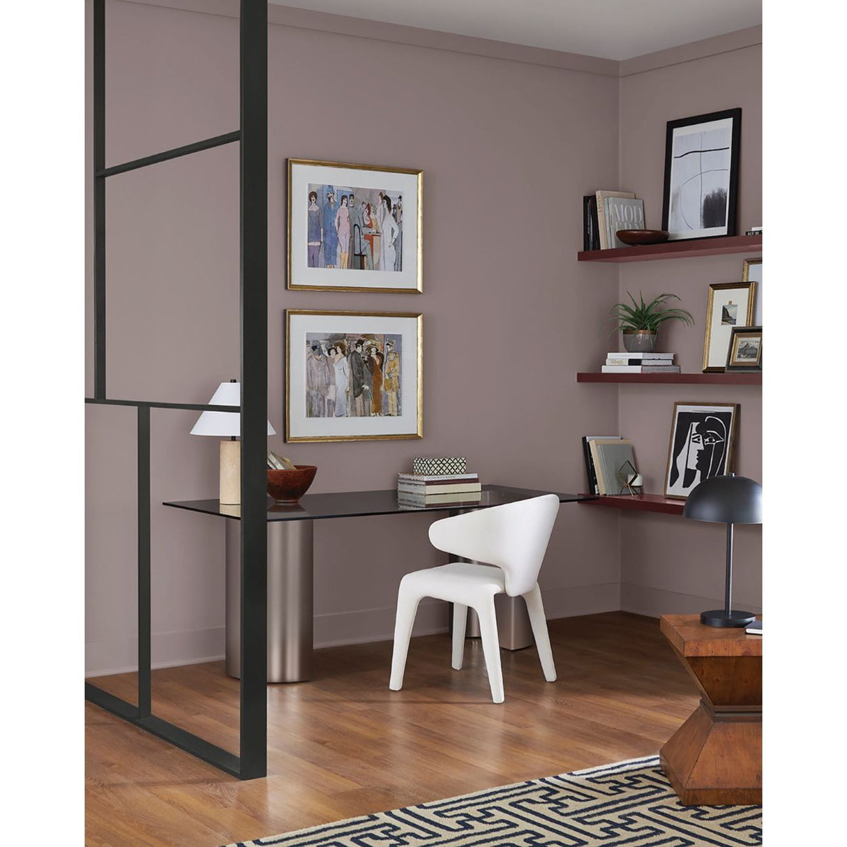 A styled home office corner with mauve-pink walls, a black desk, a white molded chair, framed artwork, floating shelves, wood flooring, and a black-and-white geometric rug.