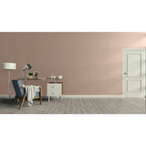 A minimalist room scene with a dusty pink wall, gray wood floor, a white door on the right, and a furnished study area on the left featuring a blue armchair, desk, lamp, and small cabinet.