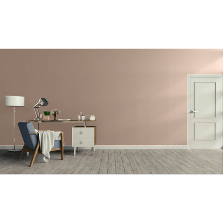 A minimalist room scene with a dusty pink wall, gray wood floor, a white door on the right, and a furnished study area on the left featuring a blue armchair, desk, lamp, and small cabinet.