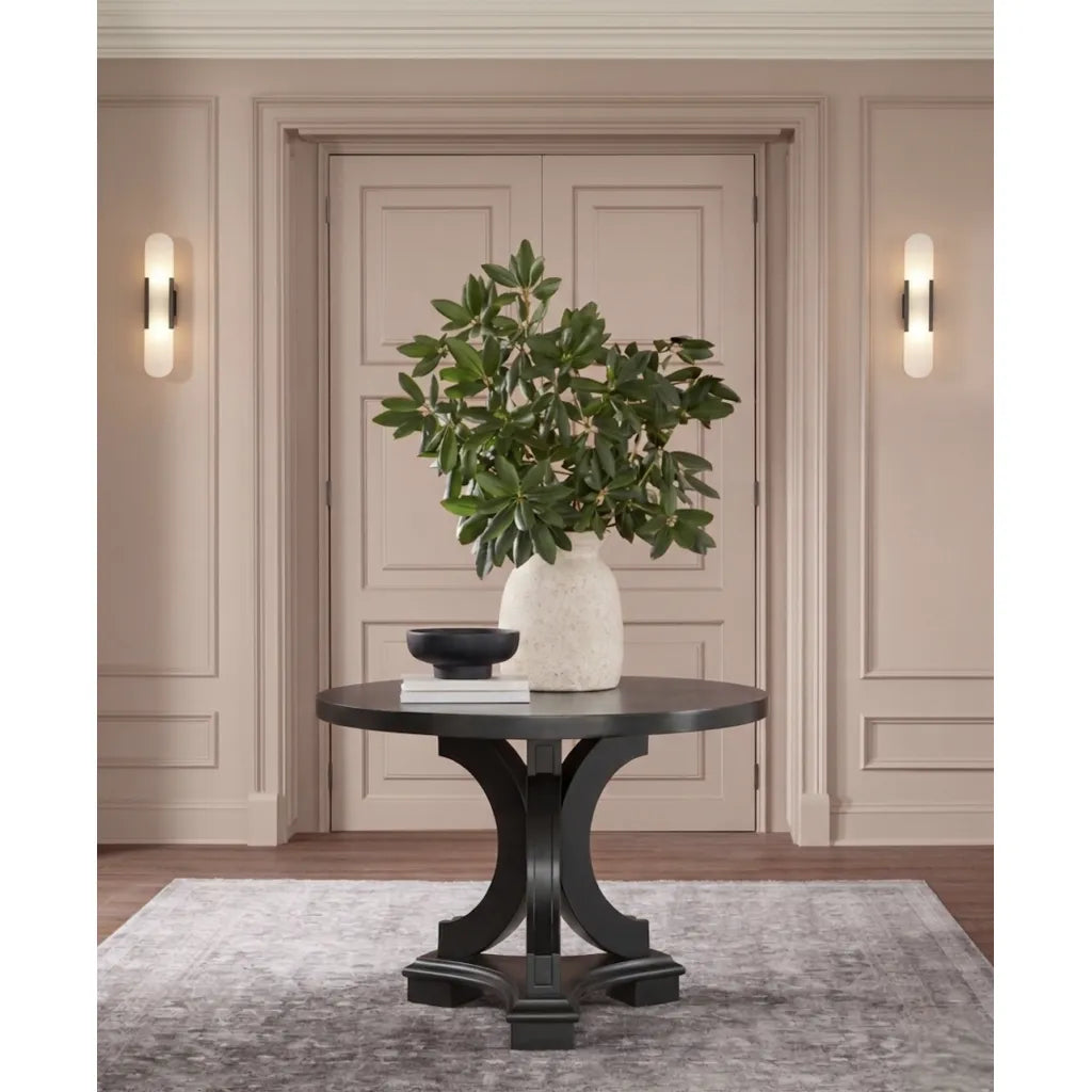 A round black console table with a sculptural curved pedestal base, styled in a warm beige entryway with a white textured vase of green foliage, a small black bowl, and books on top.
