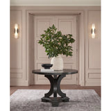 A round black console table with a sculptural curved pedestal base, styled in a warm beige entryway with a white textured vase of green foliage, a small black bowl, and books on top.