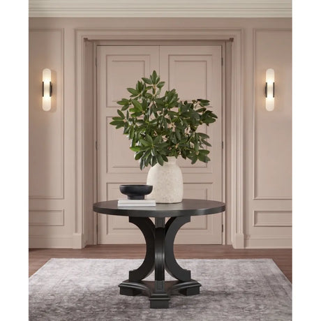 A round black console table with a sculptural curved pedestal base, styled in a warm beige entryway with a white textured vase of green foliage, a small black bowl, and books on top.