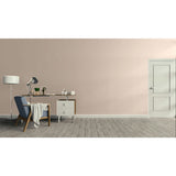 A minimalist interior room scene with a pale blush wall, gray wood floor, a blue upholstered armchair with a draped throw, a desk with a lamp and small accessories, and a white door on the right.