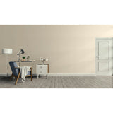 A minimalist interior room scene with a beige wall, gray wood floor, a white door on the right, and a home office setup on the left featuring a desk, lamp, chair, and small cabinet.