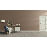 A minimalist room scene with a large warm taupe wall, light wood floor, a white door on the right, and a home office setup on the left featuring a blue armchair, desk, lamp, and small cabinet.