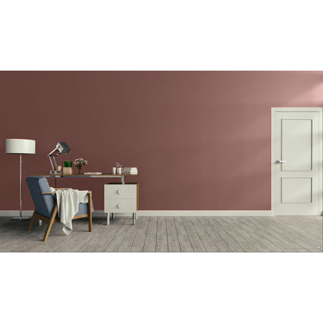 A modern interior room with a dusty rose wall, light wood floor, a white door on the right, and a home office setup on the left featuring a blue armchair, desk, lamp, and small cabinet.
