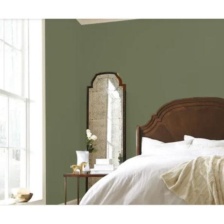 A styled bedroom corner with sage green painted walls, a wooden bed with white bedding, a tall arched mirror, and a small side table near a bright window.